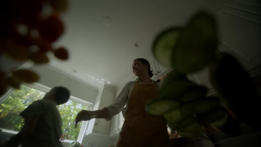 A teenager arrives in the kitchen and eats something while a woman takes a knife and cuts some food
