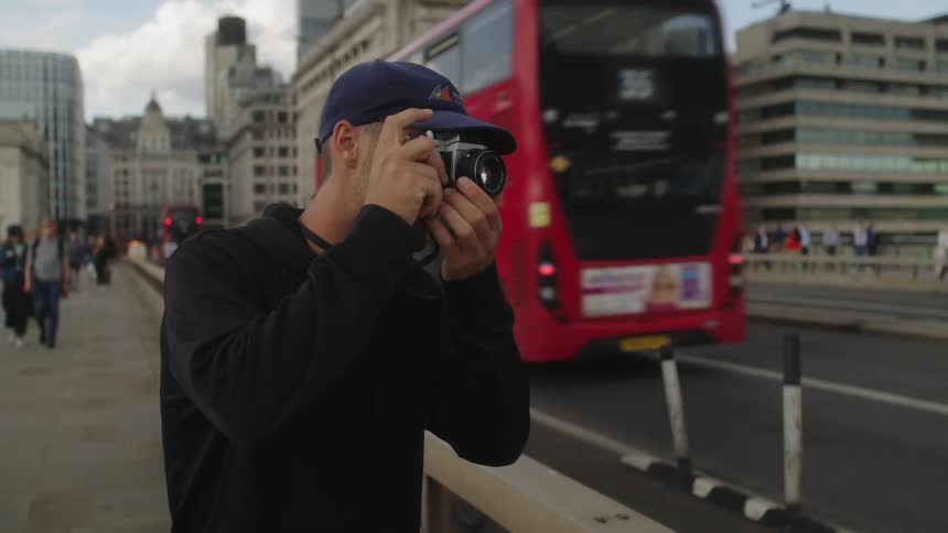 A photographer takes a photo of a double decker bus passing by on the London Bridge