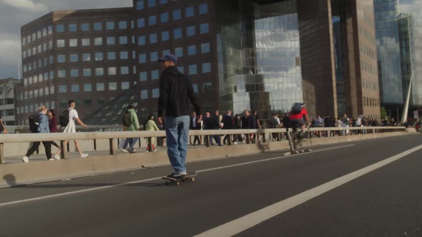 Man with cap black hoodie and jeans does tricks riding skateboard down the road next to pedestrains