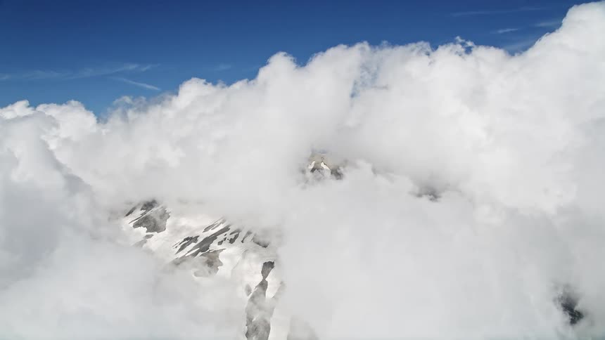 Aerial footage of Mount St Helens reveals snowy slopes and thick clouds amidst a partly cloudy sky in Washington's Cascade Mountains