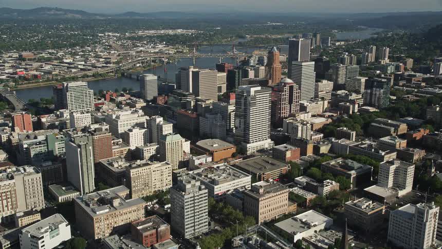 an aerial view of downtown Portland Oregon on a sunny day flying over high-rise buildings towards the majestic skyscrapers as the camera pans right showcasing the beautiful city river and surrounding nature
