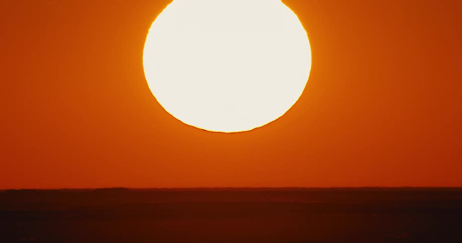 A glowing sun sets in a deep red sky as heat rises from the ground