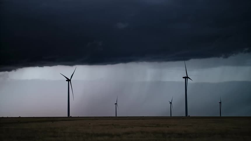 Turbines stand on a wind farm as a dark black storm cloud moves over and lightning flashes and strikes