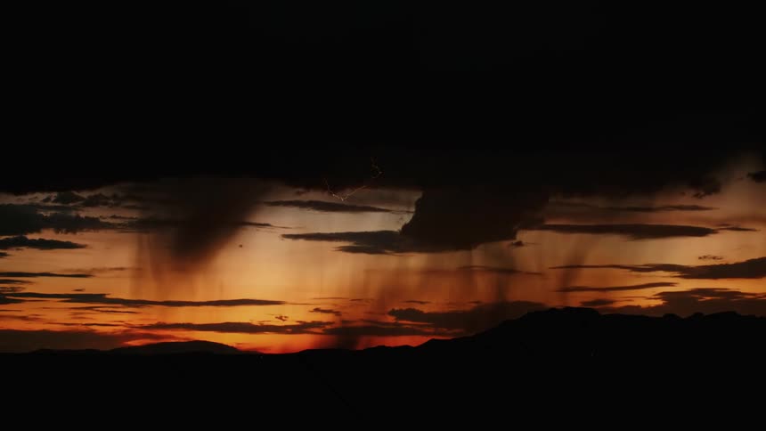 Lightning striking across the dark cloudy sky as rain pours down against an orange sunset sky