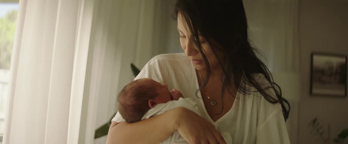 A mother holding her newborn baby swaying back and forth while standing in front of a window during the day