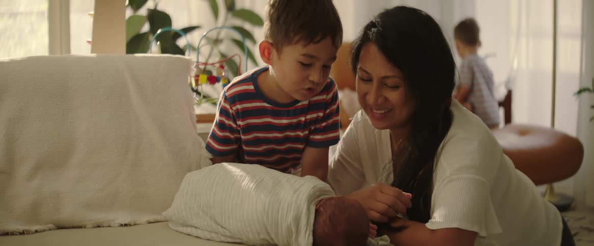 In the living room a woman kneels on the floor beside her newborn baby lying on a chair with her young son standing beside her and having a conversation in the background her other child is playing