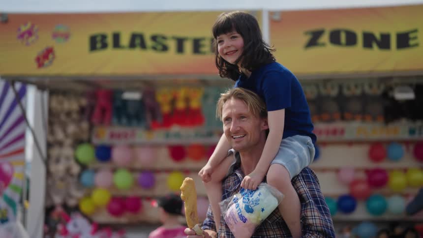 A young girl sitting on her fathers shoulders in front of a fair booth holding cotton candy in her hand as they smile and walk forward