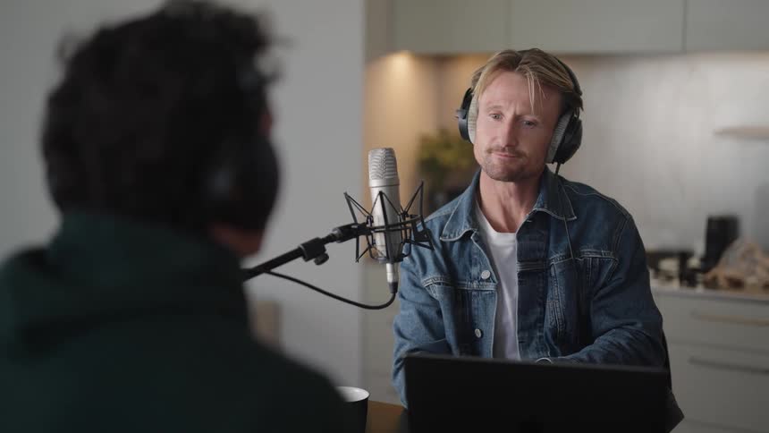 Two men sitting across from each other wearing headphones and talking into microphones indoors during the day