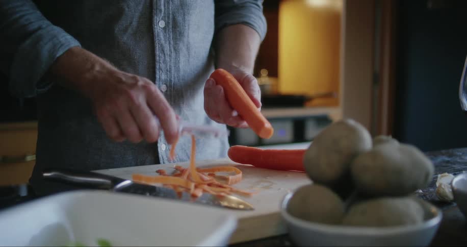 Close up of a man peeling a carrot onto a cutting board in his kitchen during the day