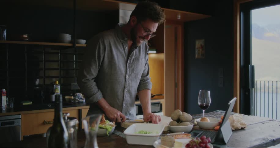 A man cooking in the kitchen while talking to someone through video call on his ipad placed on the kitchen table