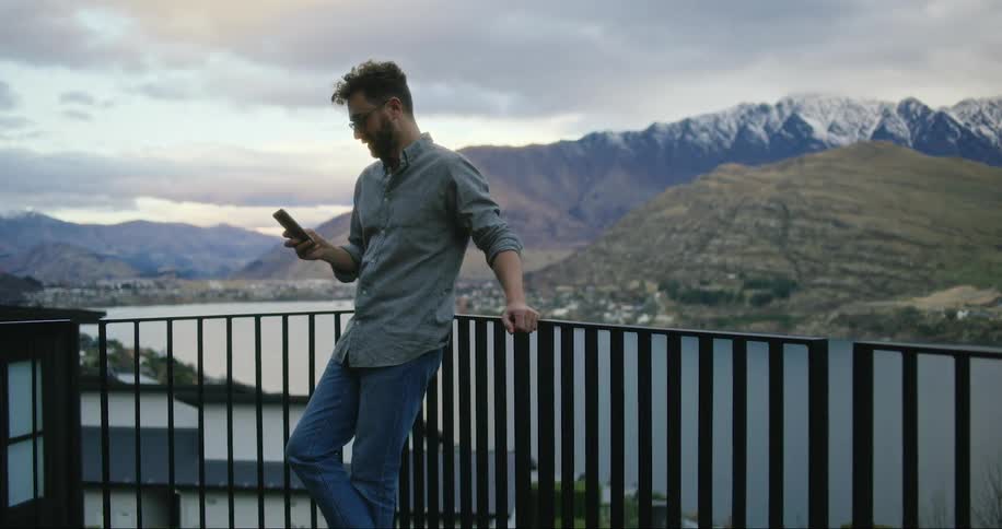 Man using his cell phone on the porch and around him mountains and lakes