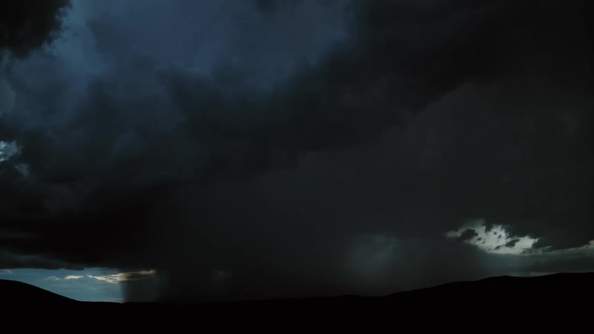 A dark supercell storm moves near mountains pouring down rain as lightning flashes inside the clouds and cracks across the sky as it strikes