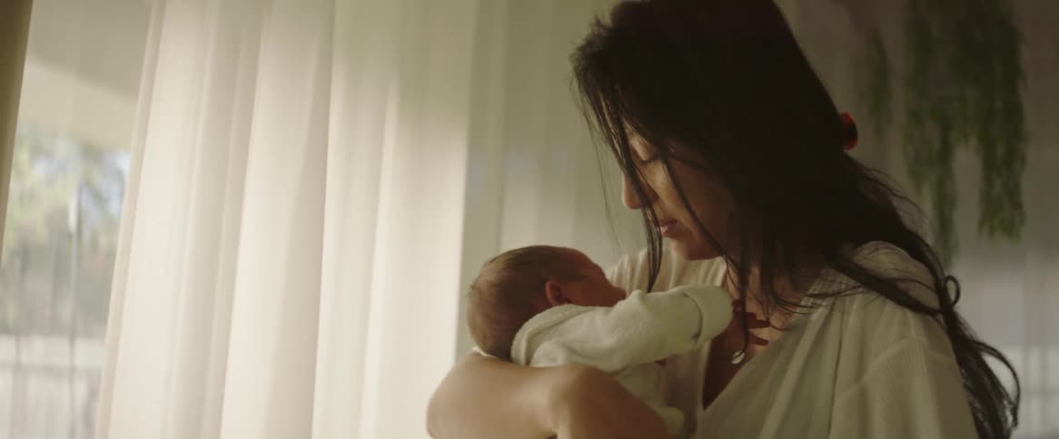 A mother holding her newborn baby swaying back and forth standing in front of a window as the sun shines through in the middle of the day