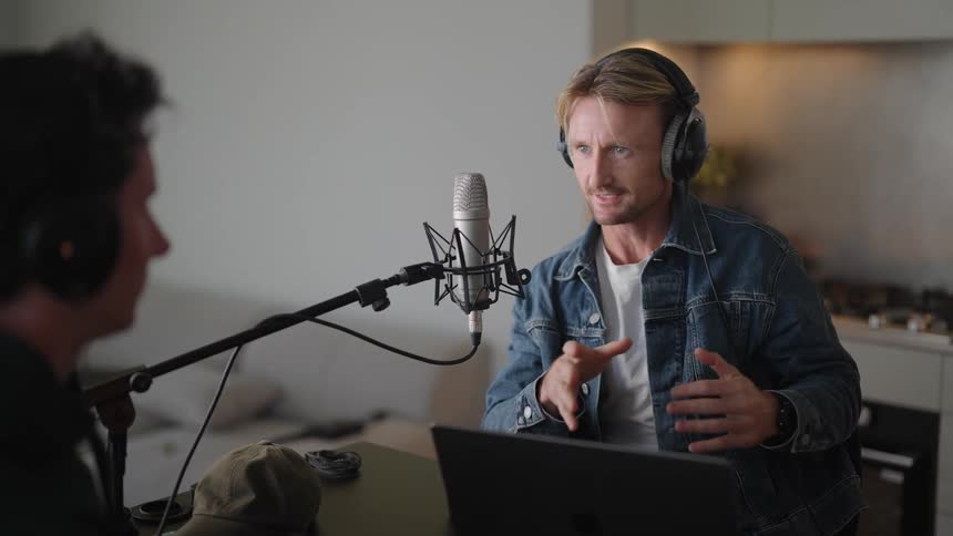 Two men sitting across from each other wearing headphones and talking into microphones in front of them as they converse