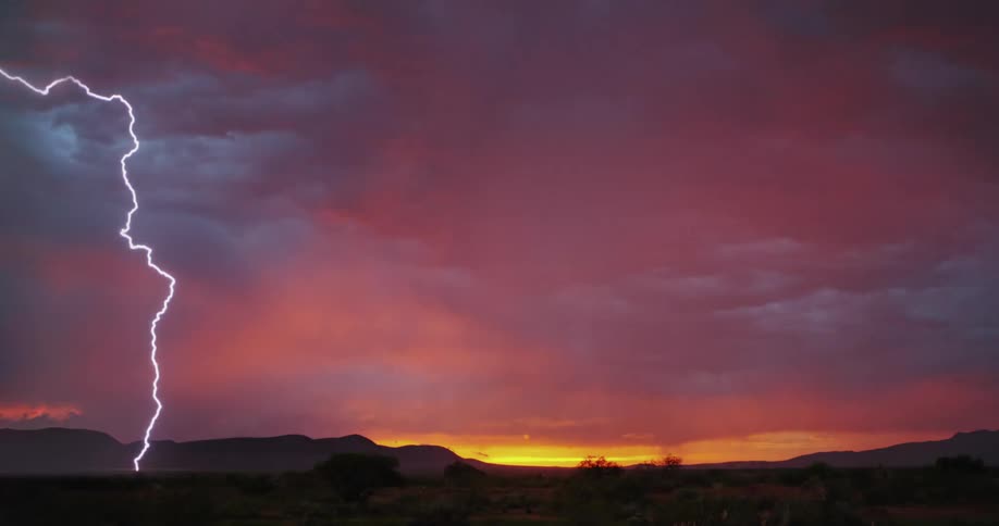 Mountains are silhouetted under a deep red sky as storm clouds move in and lightning flashes as it strikes the ground