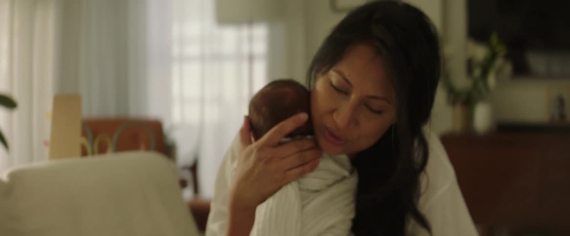 A woman picking up her infant baby from the couch and placing him beside her shoulder while sitting on the couch
