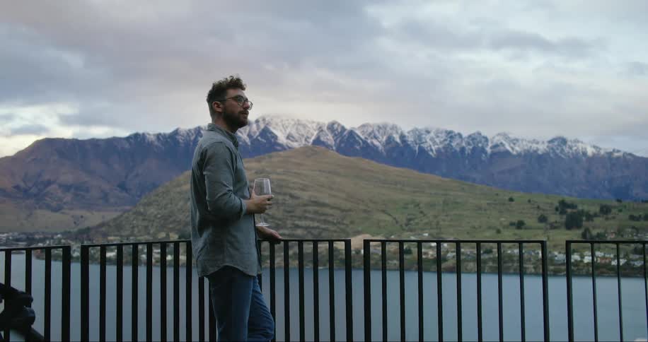A man standing outdoors on a balcony drinking wine looking out over a lake with snow covered mountains in the distance