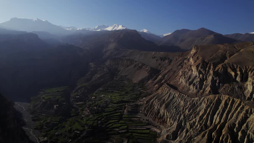 An aerial of rocky mountain ridges with a valley in the middle and snow covered peaks on the horizon