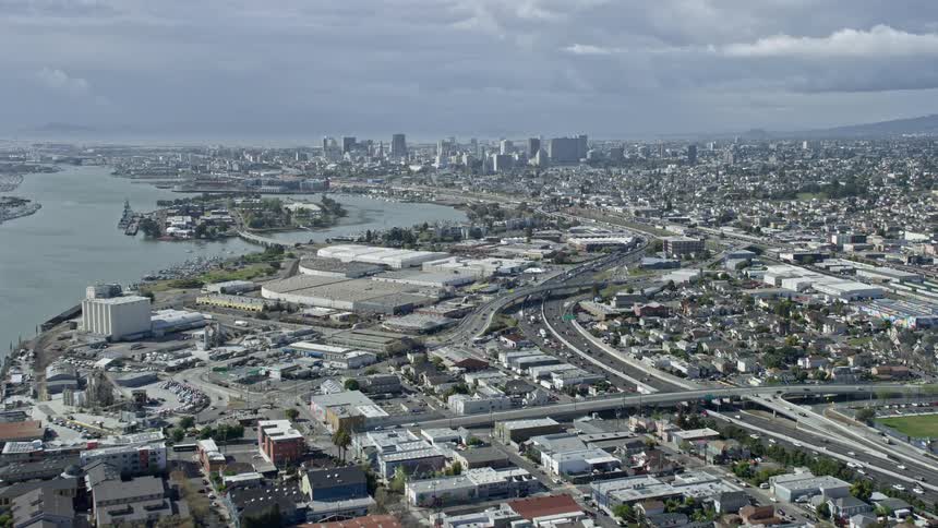  aerial footage showcasing the approaching warehouses and Downtown Oakland skyline in California with a partly cloudy sky on a summer day