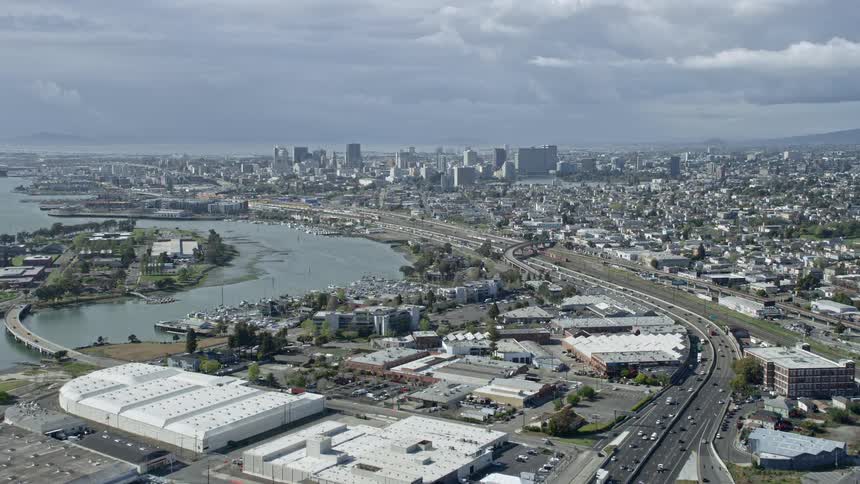 Approaching Downtown Oakland skyline in California with a busy highway winding past commercial buildings and a river showcasing the cityscape on a partly cloudy day with heavy traffic