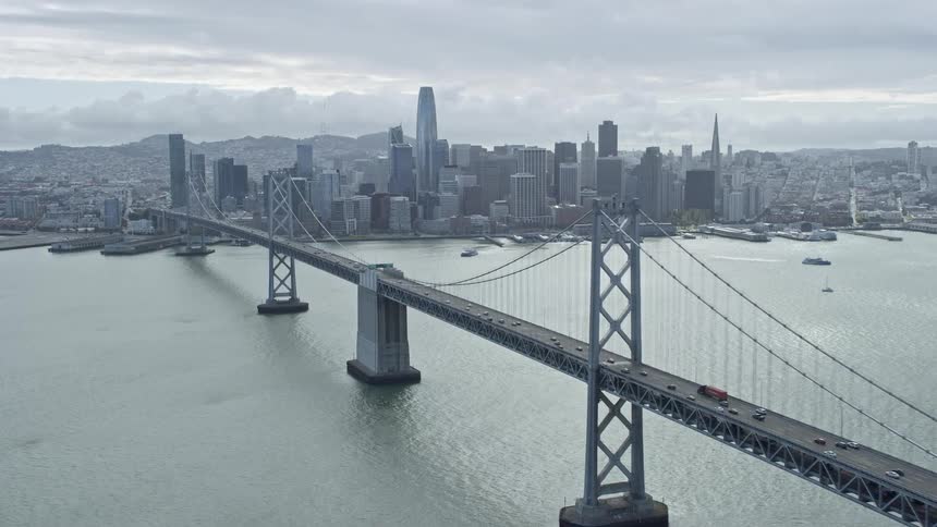  aerial footage of the Bay Bridge approaching Downtown San Francisco skyline with cloudy skies and light traffic in the background