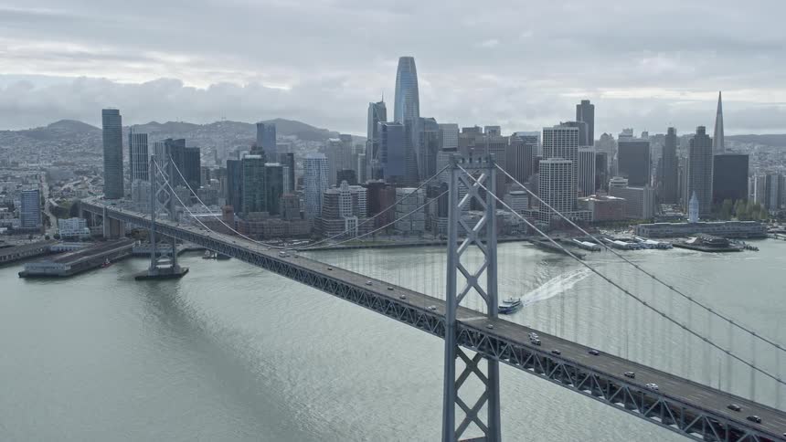  aerial footage capturing the stunning San Francisco skyline and Bay Bridge showcasing the bustling downtown area and the iconic suspension bridge in cloudy skies with light traffic