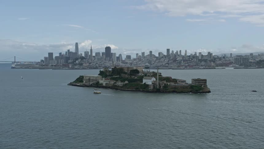 A stunning aerial view of historic Alcatraz prison orbiting above San Francisco Bay with a cloudy skyline backdrop showcasing the iconic structure and the downtown area of the city