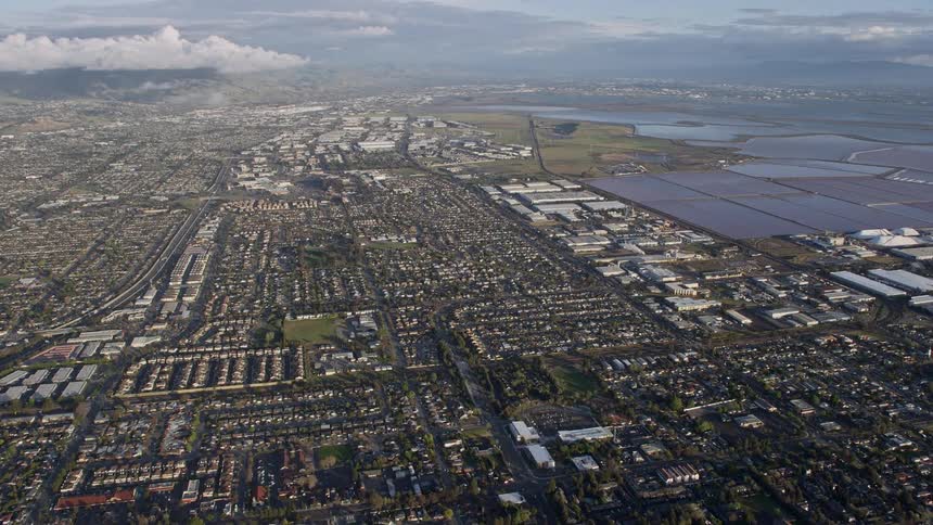  aerial footage captures the approach towards residential homes and warehouse buildings in Newark California with partly cloudy skies and a wide-angle POV