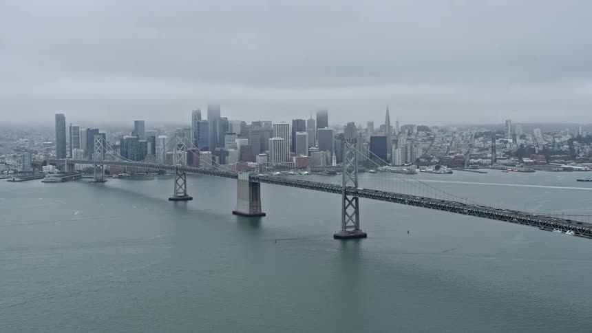  aerial footage showcasing a wide view of Downtown San Francisco and the iconic Bay Bridge from a side angle captured on a cloudy day during the spring or summer season in California