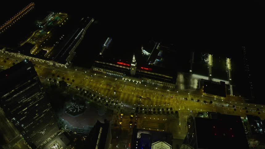 A stunningaerial shot capturing the iconic Ferry Building and the dazzling lights of Downtown San Francisco at night as the camera tilts to a bird's eye view