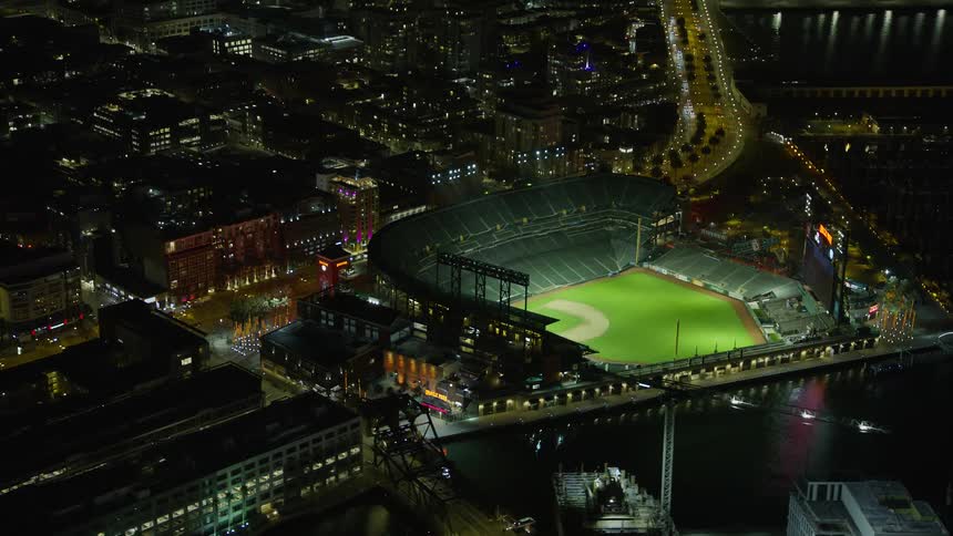 Aerial footage of Oracle Park stadium surrounded by city buildings lit up at night while circling around capturing the vibrant atmosphere of a baseball game in San Francisco California