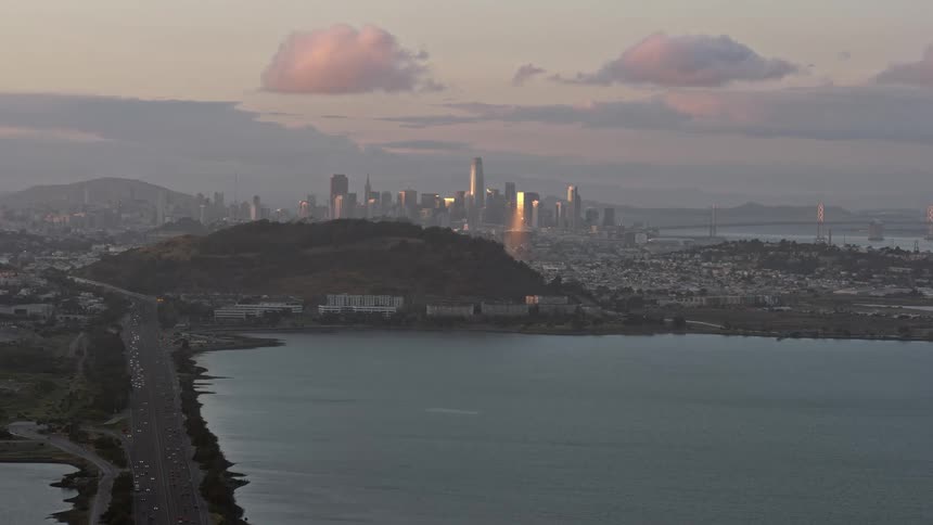 anaerial footage showcasing the stunning San Francisco skyline at sunset seen from Bayview Park in Northern California with high rises skyscrapers and downtown areas captured in wide angle as traffic approaches the city on a wide road