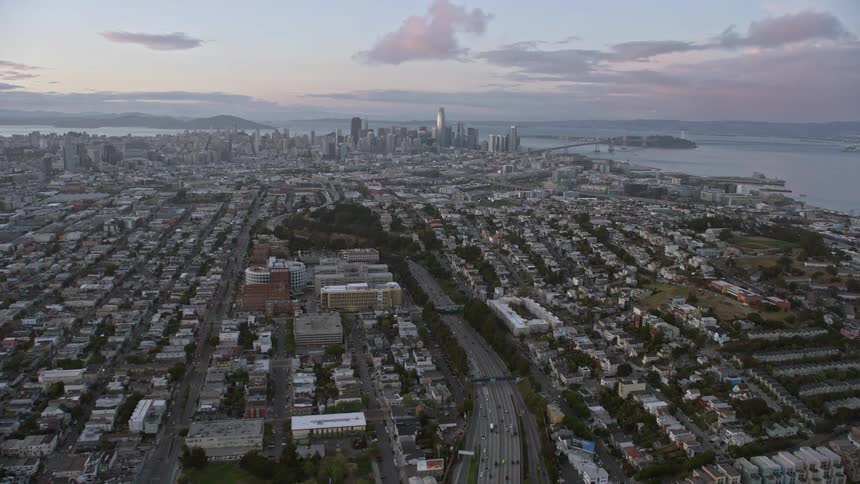 anaerial footage capturing the journey on the busy 101 freeway towards the stunning San Francisco skyline at sunset showcasing the high rise buildings downtown area and transportation in the northern California bay area with partly cloudy skies creating a