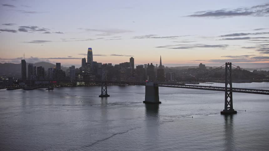  aerial footage of Downtown San Francisco skyline at twilight as the camera flies away showcasing the high rise buildings suspension bridge and partly cloudy skies capturing the beauty of the Bay Area during summer