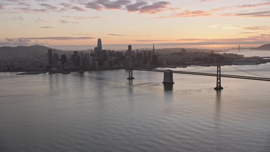 an aerial view of the Bay Bridge and the downtown San Francisco skyline at sunset showcasing the high rise buildings suspension bridge and partly cloudy skies in Northern California