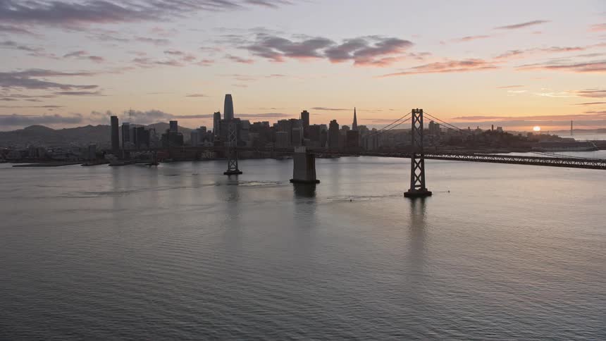 Aerial footage at sunset of flying beside the Bay Bridge toward the downtown San Francisco skyline showcasing the high rise buildings and cityscape as the sun sets over the bay area in California
