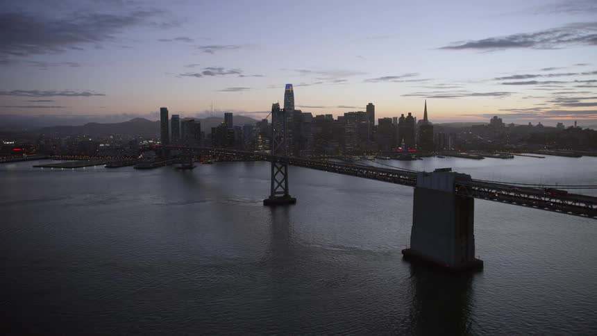  aerial footage capturing the breathtaking view of the Bay Bridge leading towards the iconic Downtown San Francisco skyline at twilight showcasing high rises skyscrapers and the stunning cityscape as partly cloudy skies create a captivating ambiance provi