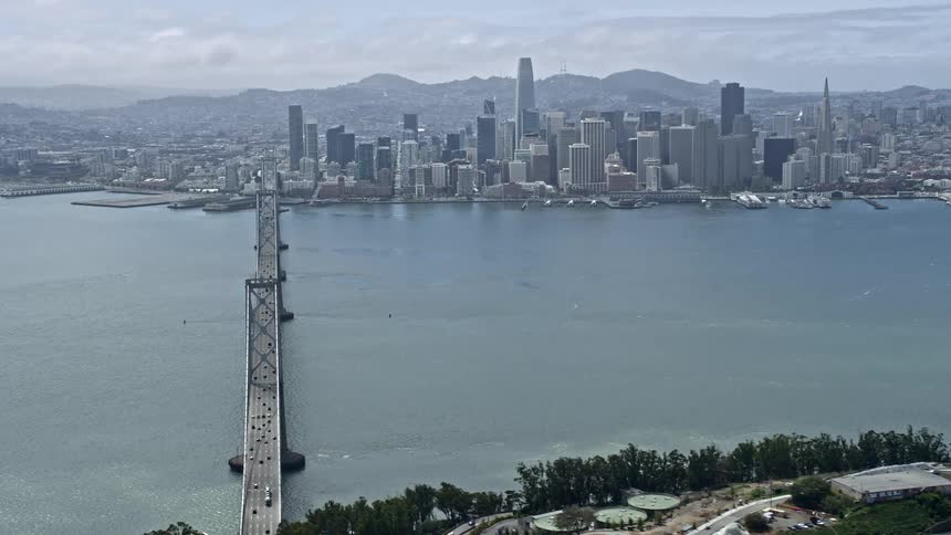 Aerial footage of San Francisco's downtown skyline and the Bay Bridge during a partly cloudy day showcasing the city's high rise buildings and the iconic suspension bridge