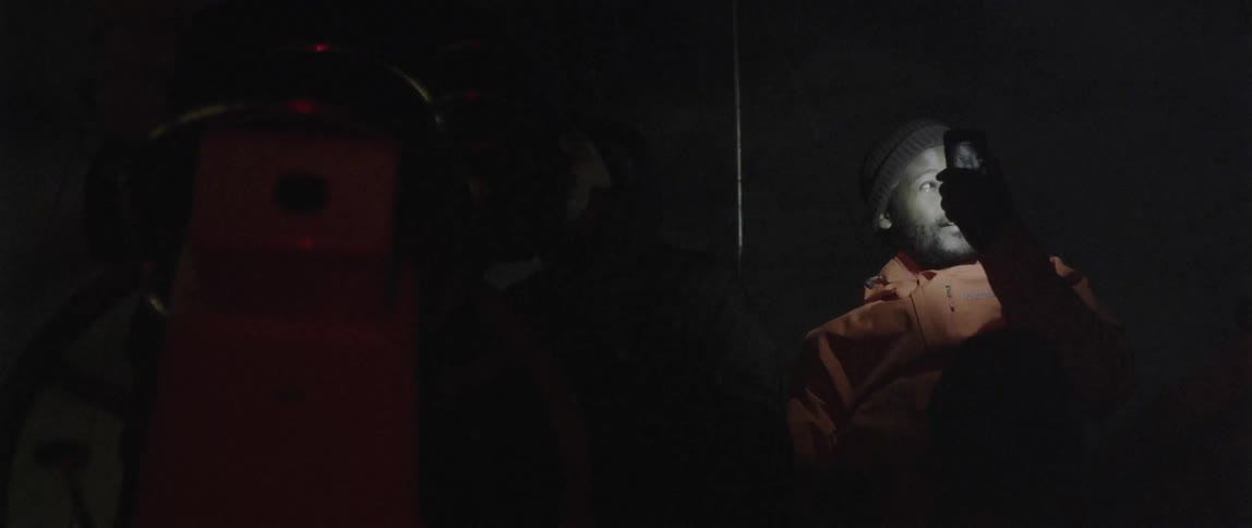 A man is filming with a cellphone as he sits in the dark with another man