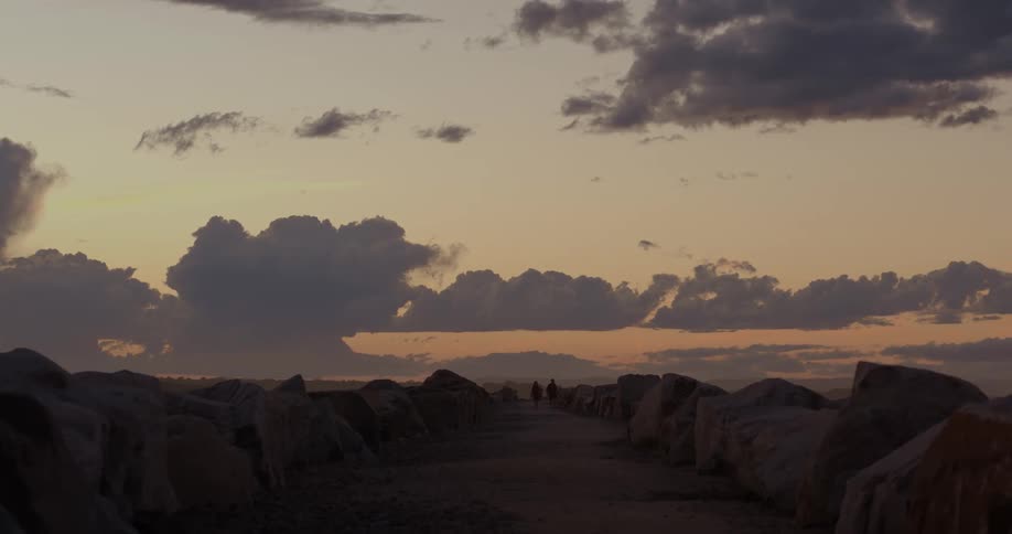 Two people walk along a path lined with large rocks under a scenic sky