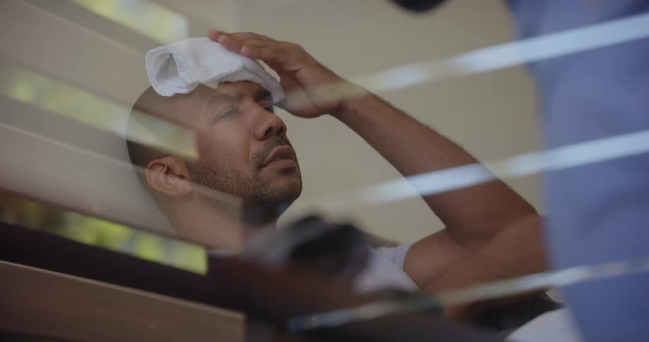 A man in pain is sitting in bed while a nurse puts a towel on his head