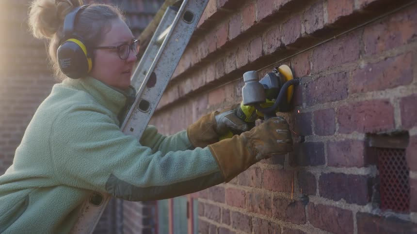 A woman standing on a ladder using a sander on a brick wall while wearing a fleece during the day