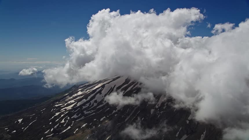 Aerial footage of Mount St Helens shows snow-covered slopes and low clouds drifting by in a partly cloudy day