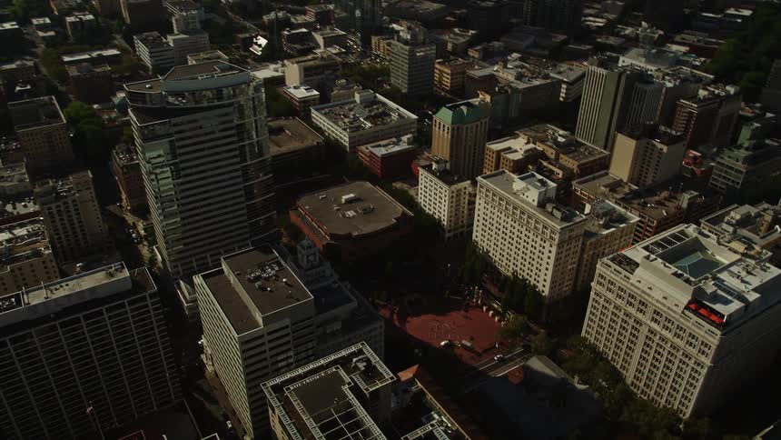Drone aerial footage of tall city buildings in downtown Portland Oregon as the sun casts shadows across the exteriors approaching the Morrison Bridge and Willamette River