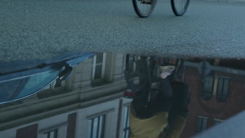 Close up of a puddle on the asphalt reflecting bikers peddling by and city buildings