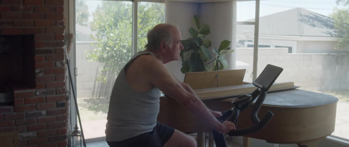 An older man pedals while atop a stationary bicycle in his home