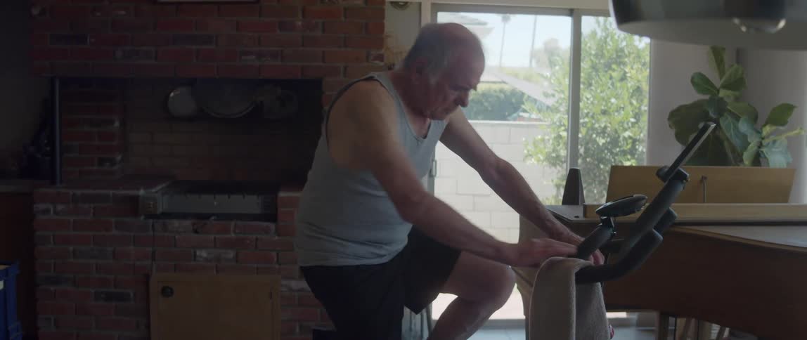 An older man adjusting himself on an exercise bike that is set up in his living room on a bright day next to a piano