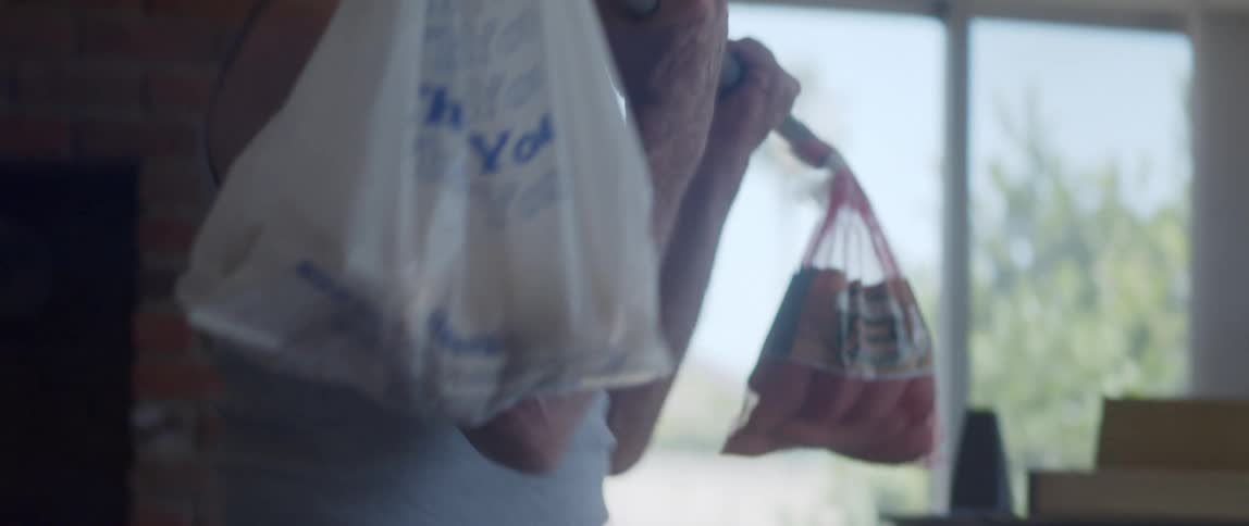 Close up of an older man lifting a weight bar with a grocery bag on one end and a sack of potatoes on the other end indoors on a bright morning