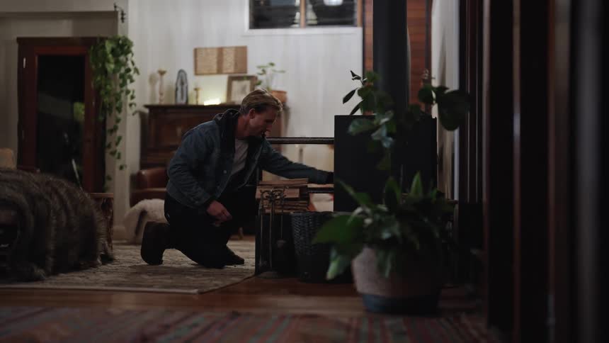 A man knelt down in front of a fire place placing sticks and wood inside while his living room is lit up by lamps in the evening
