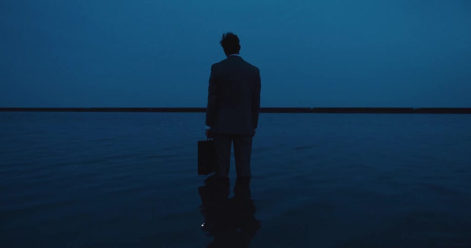 Man wearing a suit carrying a suitcase stands on the shallow coastal water at evening